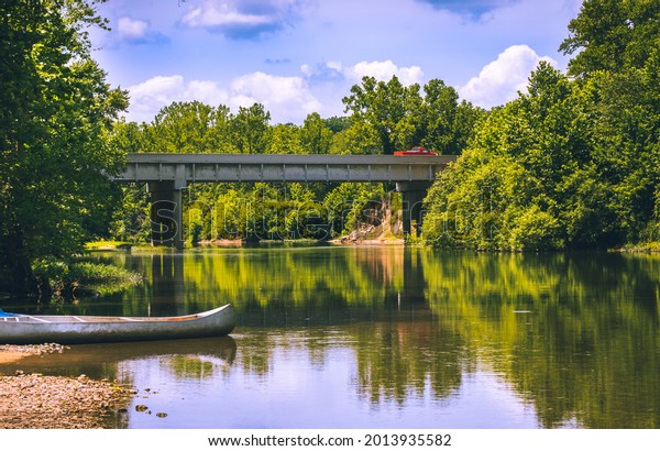Beautiful view of Midwestern river with
canoe by water edge in foreground and local highway bridge, forest
and sky in background; trees reflect in calm water

