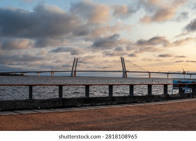 Beautiful view of a modern cable-stayed bridge spanning over a wide body of water during sunset. The image captures a dramatic sky with clouds and warm sunlight reflecting on the water, highlighting the elegant architecture of the bridge.
