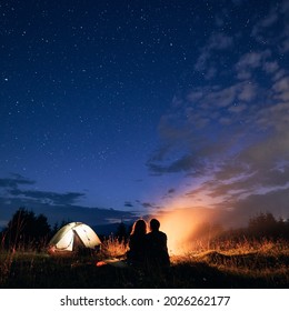 Beautiful view of night starry sky over grassy hill with illuminated camp tent, campfire and hikers. Back view of couple tourists having a rest near bonfire. Concept of night camping, relationship. - Powered by Shutterstock