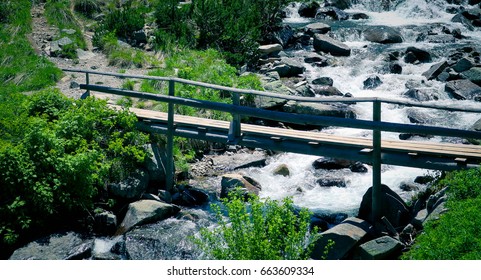 Beautiful View On The High Green Mountains Peaks And A Wooden Old Bridge Over The River, Blue Sky Background. Mountain Hiking Paradise Landscape, A Stream Flows Down The Rocks, No People.