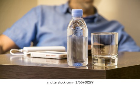 A Beautiful View Of A Plastic Small Bottle With Clear Water Or A Fresh Drink, Standing Against The Backdrop Of A Sitting Man In A Dark Room.