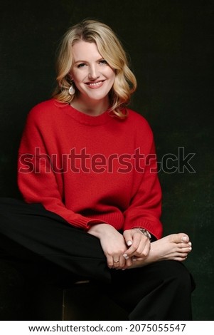 beautiful view of a smiling blonde girl in a red sweater sitting on a drawer and with her legs crossed over her knee