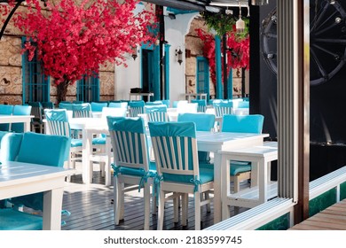 Beautiful View Of Summer Street Cafe With White-blue Tables And Chairs. Open Air Restaurant On Background Of Red Bougainvillea On The Wall With No People. Selective Focus.