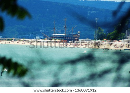 Beautiful view of the white beaches in Varna, Bulgaria, Europe. Scenery of sea waters, sunbathing people on the beach, sun umbrellas and a ship behind them.  The picture is in a frame of tree branches