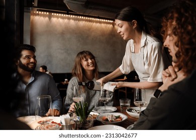 Beautiful waitress wearing apron pouring wine into glasses while serving group of young friends in restaurant