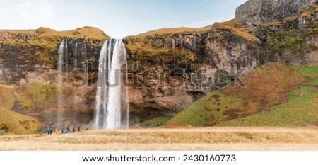 Beautiful waterfall flowing through rocky mountains covered with green grass in sunny daylight and enjoying visitors on ground in countryside. The Golden Circle, Iceland