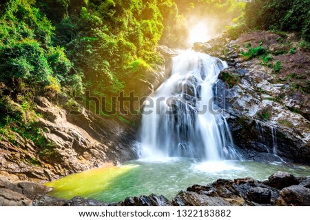 Beautiful waterfall at the mountain with blue sky and white cumulus clouds. Waterfall in tropical green tree forest. Waterfall is flowing in jungle. Nature abstract background. Granite rock mountain. 