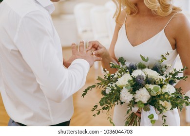 Beautiful Wedding Celebration. A Pregnant Blonde Bride In Simple White Dress With A Bouquet Of Light Flowers And Green Twigs Puts Engagement Ring Her Groom In Gray Trousers And A White Shirt.