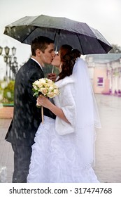 Beautiful Wedding Couple Kissing In The Rain. Bride And Groom Walking On The Street Under An Umbrella On A Rainy Day