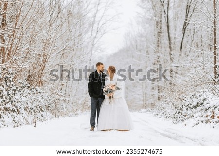 Beautiful wedding couple walking in winter snowy forest, woman in white dress and mink fur coat, bearded man in black coat