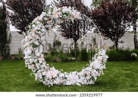 A beautiful wedding round arch, decorated with flowers, electric hanging lamps, stands on the green grass of a lawn in a garden, park. Festive photography in nature.