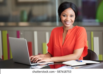 Beautiful Well Dressed Young Black Businesswoman Looking At The Camera While Sitting At Her Desk In Her Colourful Office Environment, Busy Typing On Her Modern Laptop Computer.