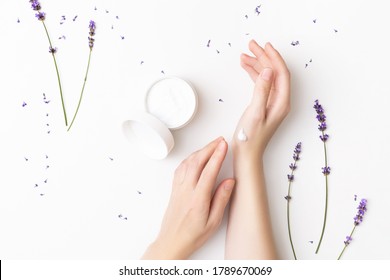 Beautiful Well-conditioned Female Hands Moisturizing Cream Surrounded With Lavender Flowers On The White Background. Hand And Body Care Concept