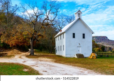 A beautiful white Baptist church stands alongside a dirt road in front of Arkansas trees full of autumn foliage in colors such as red, green, yellow, gold and brown. 