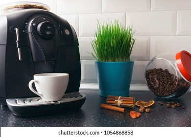 Beautiful white kitchen in an apartment with a coffee machine, a white cup, spices, a glass jar with coffee beans and a green flower in a blue pot. Black surface of the kitchen table