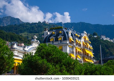 Beautiful white multi-storey building with balconies and yellow tents awnings on windows surrounded by green trees in front of mountains Alps, Swiss Riviera, Vaud canton, Switzerland