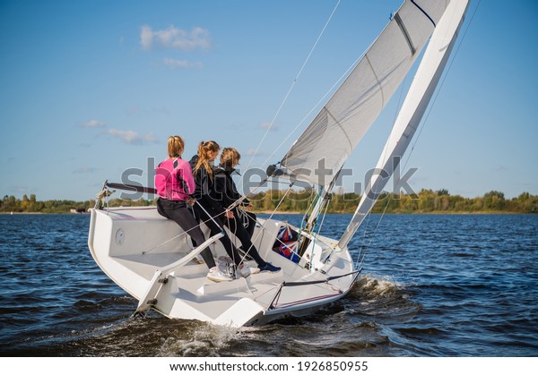 A beautiful white racing single-masted yacht is\
sailing against a beautiful river landscape with a blue sky. A man\
and two girls are on board