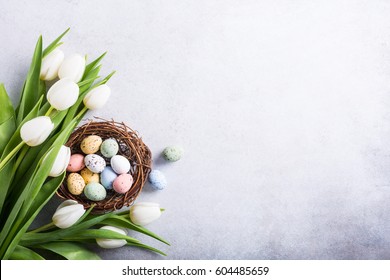 Beautiful white tulips with colorful quail eggs in nest on light gray stone background. Spring and Easter holiday concept with copy space.