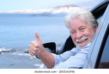 Beautiful White-haired Senior Man Inside The Car Looking At Camera With Thumb Up. Smiling White Haired Elderly People Enjoying Freedom And Retirement. Seascape And Blue Sky