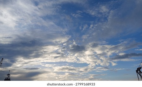 A beautiful wide-angle view of a dramatic sky during golden hour. Fluffy white and gray clouds spread across the vibrant blue sky, with soft sunlight breaking through. Industrial crane and ship mast silhouettes are visible at the bottom frame, creating a contrast between nature and industry. Perfect for backgrounds, weather concepts, freedom, hope, and industrial themes.