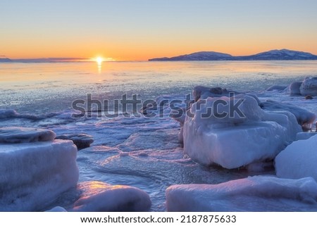 Beautiful winter arctic seascape. The sun sets over the horizon. Ice-covered seashore and unfrozen water. Large ice floes on the shore. Sunset landscape with sea coast. Cold weather. Northern nature.