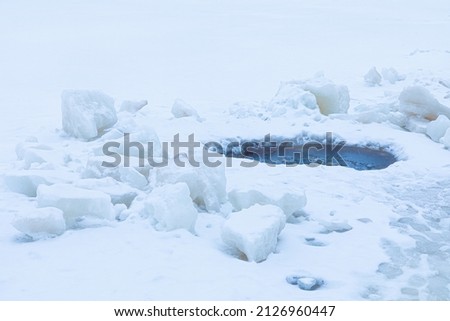 Beautiful winter landscape of bright, vivid and colorful ice hole among snow surface of lake, river or pound during snowfall or blizzard