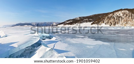 Beautiful winter landscape of frozen Lake Baikal with ice hummocks and ice breaks. The Great Baikal Trail of tourists is visible on the coastal hill. Panoramic view. Natural background. Ice travel