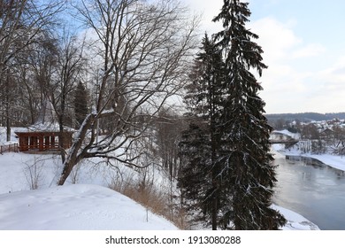 Beautiful winter landscape. Picturesque trees on the hillside. Tall spruce and deciduous tree with a spreading crown. A calm river, snowdrifts and a wooden gazebo. Wonderful nature in the park.