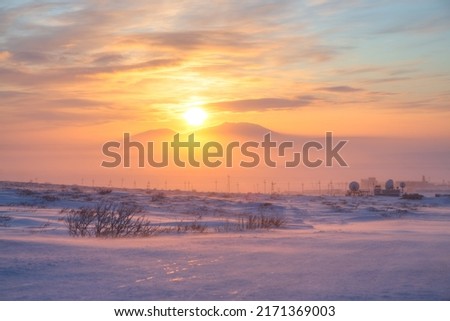 Beautiful winter sunset in the Arctic. View of the snowy tundra and the setting sun over the mountains. Cold frosty winter weather. Blowing snow. Satellite dishes in the tundra. Chukotka, Russia.