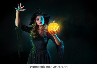 beautiful witch with hat conjures over jack-o-lantern pumpkin. Halloween party, witches ' Sabbath. Young beautiful brunette in witch costume, photo with art elements, magic smoke and grunge background
