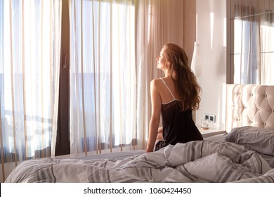 The beautiful woman after awakening sitting on a bed in the bedroom and rejoicing to sunrise.