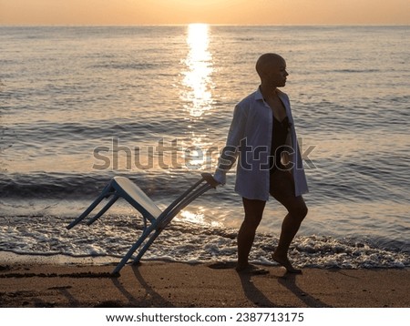 beautiful woman with alopecia relaxing on seashore,sitting or dragging chair in water,sunset or sunrise time. girl with no hair is walking on beach,dancing with white shirt as with a man. crazy fun 