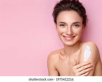 Beautiful woman applying body lotion on her shoulder. Photo of smiling woman with perfect makeup on pink background. Beauty concept