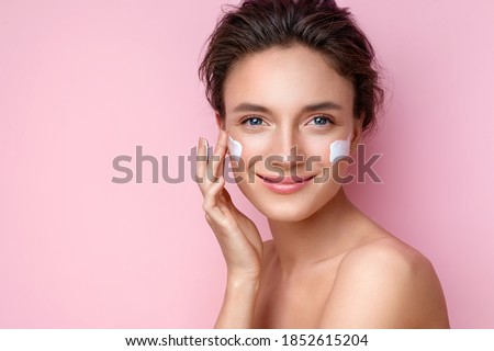 Beautiful woman applying moisturizer cream on her face. Photo of smiling woman with perfect makeup on pink background. Beauty concept