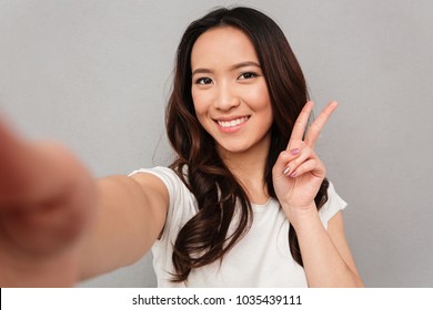 Beautiful Woman With Asian Appearance Taking Selfie And Showing Victory Sign With Perfect Smile Isolated Over Gray Background