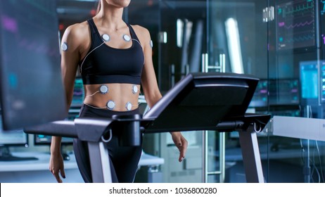 Beautiful Woman Athlete With Electrodes Attached To Her, Running On A Treadmill In A Sports Science Laboratory. In The Background Lab With Monitors Showing EKG Data.