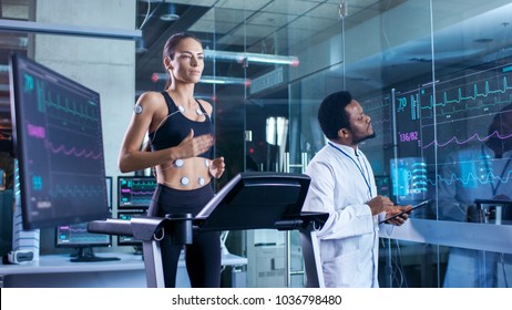 Beautiful Woman Athlete Runs on a Treadmill with Electrodes Attached to Her Body, Physician Uses Tablet Computer and Controls EKG Data Showing on Laboratory Monitors.