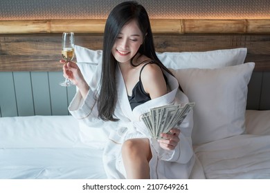 Beautiful Woman In Bathrobe With Glass Of Champagne In Bedroom Holding Cash Money On Hand. Young Rich Asian Woman With Glasses Of Champagne And Holding Money. 