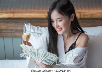 Beautiful Woman In Bathrobe With Glass Of Champagne In Bedroom Holding Cash Money On Hand. Young Rich Asian Woman With Glasses Of Champagne And Holding Money. 