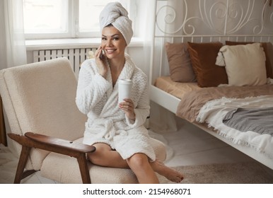 Beautiful woman in bathrobe and towel after shower talking on phone and drinking coffee at bedroom