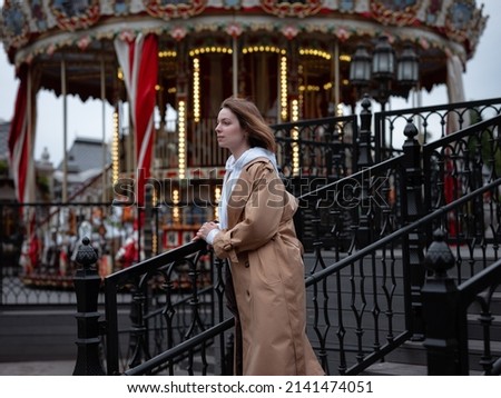 Beautiful woman in beautiful beige coat walk in autumn cloudy Moscow, enjoys and has fun on carousel. Street style. Travel to Russia concept.