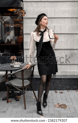 beautiful woman with beret is drinking cappuccino in outdoor cafe autumn fall time. girl doing kissing gesture, smiling, having good time. Parisian style, old street black and white colors