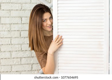 Beautiful Woman With Big Blue Eyes In A Strict Dress Looking Out From Behind The Wooden Door Of Dressing Room. Brunette With Light-colored Hair.