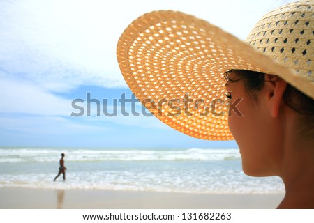 Beautiful woman with big straw hat looking at a man walking on the beach