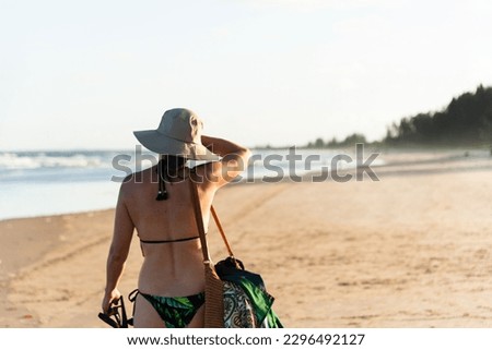 Beautiful woman in bikini, holding hat on head, walking on sandy beach. Guaibim, Valenca, Bahia.