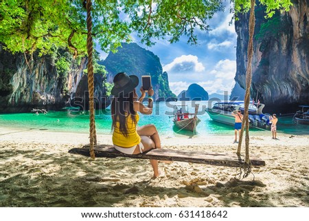Beautiful woman in bikini sitting wooden swing under tree using tablet on tropical beach, Koh Lading island, Andaman sea, Krabi province, Thailand