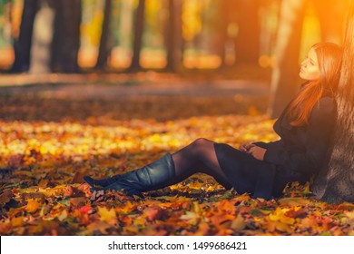 Beautiful woman in black coat and leather boots sitting under tree in autumn park with fallen leaves. Bright young long haired brunette girl relax in park with red yellow maple leaves in fall season
