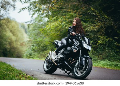 Beautiful Woman In A Black Leather Jacket Sitting On A Black Sports Motorcycle On The Road In The Woods