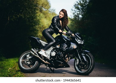 Beautiful Woman In A Black Leather Jacket Sitting On A Black Sports Motorcycle On The Road In The Woods