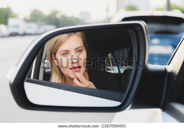 Beautiful woman with blond hair driving a car paints\
her lips with lipstick. The woman driving making make-up.\
Reflection of a woman in the rearview mirror. Photo of a successful\
woman in a car.